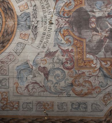 Zdjęcie nr 13: Polichromia umieszczona na stropie nawy głównej. W polu środkowym scena Wniebowzięcia Najświętszej Marii Panny umieszczona w kwadratowej ramie o bokach zamkniętych łukami nadwieszonymi. Rama profilowana, zdobiona rzędem pojedynczych liści. Matka Boska unosząca się na obłokach z szeroko rozłożonymi na boki rękami, prawą wzniesioną do góry. Twarz owalna z długim nosem, silnie zaciśniętymi ustami i dużymi oczami, skierowanymi ku górze. Włosy jasne, kręcone, schowane pod żółtym welonem, którego jeden koniec rozwiany jest wzdłuż prawej ręki. Matka Boska ubrana jest w szeroką, różową suknię, przewiązaną w talii pomarańczową tasiemką oraz błękitny płaszcz, założony za lewe ramię i rozwiany z tyłu. Nad głową Matki Boskiej aniołki unoszą insygnia królewskie: koronę, berło, a także gałązkę białej lilii. Poniżej grupy aniołów podtrzymujące obłoki. Dokoła ramy bujne sploty różnokolorowego akantu przeplecionego karbowaną wstęgą, owijające również ramę ze sceną Wniebowzięcia. W dolnej części ukazane są ponadto dwa anioły podtrzymujące białą wstęgę z napisem: „ASSVMPTA EST MARIA IN COELVM, GAV, / DENT ANGELI, LAVDANTES BENEDICVNT / DOMINVM.” Z kolei z drugiej strony stropu umieszczony jest owalny medalion, obwiedziony liśćmi wawrzynu, w polu którego umieszczone jest czerwone godło zakonu Bożogrobowców oraz małe uskrzydlone główki aniołków. Powyżej na białej wstędze umieszczony jest napis: „SVB HOC SIGNO.”, a poniżej: „RESTAVRATVM A(NN)O (DOMINI) 1714 / Die 14 Aug(st)”. Całość stropu obwiedziona jest dookoła pasem przeważnie prostokątnych kasetonów, wypełnionych różnokolorowymi liśćmi akantu, a także od zewnątrz szerokim pasem dekoracyjnym z brązowych liści wawrzynu. Pasy na bokach wyłamane półkolistymi wypustkami, w których umieszczone są duże muszle palmetowe. Kolorystyka stonowana, jasna z przewagą błękitu i czerwieni.