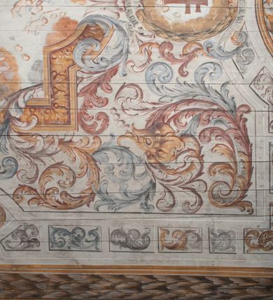 Zdjęcie nr 14: Polichromia umieszczona na stropie nawy głównej. W polu środkowym scena Wniebowzięcia Najświętszej Marii Panny umieszczona w kwadratowej ramie o bokach zamkniętych łukami nadwieszonymi. Rama profilowana, zdobiona rzędem pojedynczych liści. Matka Boska unosząca się na obłokach z szeroko rozłożonymi na boki rękami, prawą wzniesioną do góry. Twarz owalna z długim nosem, silnie zaciśniętymi ustami i dużymi oczami, skierowanymi ku górze. Włosy jasne, kręcone, schowane pod żółtym welonem, którego jeden koniec rozwiany jest wzdłuż prawej ręki. Matka Boska ubrana jest w szeroką, różową suknię, przewiązaną w talii pomarańczową tasiemką oraz błękitny płaszcz, założony za lewe ramię i rozwiany z tyłu. Nad głową Matki Boskiej aniołki unoszą insygnia królewskie: koronę, berło, a także gałązkę białej lilii. Poniżej grupy aniołów podtrzymujące obłoki. Dokoła ramy bujne sploty różnokolorowego akantu przeplecionego karbowaną wstęgą, owijające również ramę ze sceną Wniebowzięcia. W dolnej części ukazane są ponadto dwa anioły podtrzymujące białą wstęgę z napisem: „ASSVMPTA EST MARIA IN COELVM, GAV, / DENT ANGELI, LAVDANTES BENEDICVNT / DOMINVM.” Z kolei z drugiej strony stropu umieszczony jest owalny medalion, obwiedziony liśćmi wawrzynu, w polu którego umieszczone jest czerwone godło zakonu Bożogrobowców oraz małe uskrzydlone główki aniołków. Powyżej na białej wstędze umieszczony jest napis: „SVB HOC SIGNO.”, a poniżej: „RESTAVRATVM A(NN)O (DOMINI) 1714 / Die 14 Aug(st)”. Całość stropu obwiedziona jest dookoła pasem przeważnie prostokątnych kasetonów, wypełnionych różnokolorowymi liśćmi akantu, a także od zewnątrz szerokim pasem dekoracyjnym z brązowych liści wawrzynu. Pasy na bokach wyłamane półkolistymi wypustkami, w których umieszczone są duże muszle palmetowe. Kolorystyka stonowana, jasna z przewagą błękitu i czerwieni.