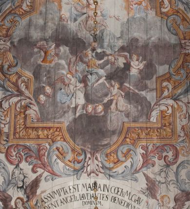 Zdjęcie nr 1: Polichromia umieszczona na stropie nawy głównej. W polu środkowym scena Wniebowzięcia Najświętszej Marii Panny umieszczona w kwadratowej ramie o bokach zamkniętych łukami nadwieszonymi. Rama profilowana, zdobiona rzędem pojedynczych liści. Matka Boska unosząca się na obłokach z szeroko rozłożonymi na boki rękami, prawą wzniesioną do góry. Twarz owalna z długim nosem, silnie zaciśniętymi ustami i dużymi oczami, skierowanymi ku górze. Włosy jasne, kręcone, schowane pod żółtym welonem, którego jeden koniec rozwiany jest wzdłuż prawej ręki. Matka Boska ubrana jest w szeroką, różową suknię, przewiązaną w talii pomarańczową tasiemką oraz błękitny płaszcz, założony za lewe ramię i rozwiany z tyłu. Nad głową Matki Boskiej aniołki unoszą insygnia królewskie: koronę, berło, a także gałązkę białej lilii. Poniżej grupy aniołów podtrzymujące obłoki. Dokoła ramy bujne sploty różnokolorowego akantu przeplecionego karbowaną wstęgą, owijające również ramę ze sceną Wniebowzięcia. W dolnej części ukazane są ponadto dwa anioły podtrzymujące białą wstęgę z napisem: „ASSVMPTA EST MARIA IN COELVM, GAV, / DENT ANGELI, LAVDANTES BENEDICVNT / DOMINVM.” Z kolei z drugiej strony stropu umieszczony jest owalny medalion, obwiedziony liśćmi wawrzynu, w polu którego umieszczone jest czerwone godło zakonu Bożogrobowców oraz małe uskrzydlone główki aniołków. Powyżej na białej wstędze umieszczony jest napis: „SVB HOC SIGNO.”, a poniżej: „RESTAVRATVM A(NN)O (DOMINI) 1714 / Die 14 Aug(st)”. Całość stropu obwiedziona jest dookoła pasem przeważnie prostokątnych kasetonów, wypełnionych różnokolorowymi liśćmi akantu, a także od zewnątrz szerokim pasem dekoracyjnym z brązowych liści wawrzynu. Pasy na bokach wyłamane półkolistymi wypustkami, w których umieszczone są duże muszle palmetowe. Kolorystyka stonowana, jasna z przewagą błękitu i czerwieni.