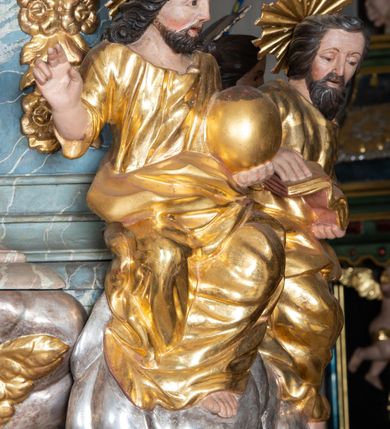 Zdjęcie nr 11: Ambona przyścienna, stojąca, z wejściem w postaci schodów z balustradą od strony prezbiterium. Kosz na planie ściętego z dwóch stron kwadratu, ustawiony pod kątem względem zaplecka, na profilowanym cokole, z podwieszonym u spodu zwężającym się w dół elementem podtrzymywanym przez figurę anioła. Dwie frontowe ścianki podzielone po środku pilastrami dźwigającymi pełne belkowanie o silnie wysuniętym gzymsie stanowiącym parapet kosza. Trzony pilastrów oraz pola pomiędzy nimi dekorowane są roślinnymi zwisami z muszlami. Na cokole kosza znajdują się siedzące na obłokach figury czterech ewangelistów z otwartymi księgami oraz na froncie figura Chrystusa trzymającego kulę. Podwieszony element pomiędzy siedzącymi figurami dekorowany jest obłokami i uskrzydlonymi główkami anielskimi. Balustrada schodów ozdobiona ornamentem roślinnym, zakończona półfilarem, na którym stoi figurka św. Jana Nepomucena. Zaplecek wąski, w formie stojącego prostokąta, z płyciną w polu ujętą profilowaną ramą w formie stojącego prostokąta zamkniętego łukiem segmentowym nadwieszonym z obrazem Chrystusa Dobrego Pasterza. Płycina flankowana rzędami kampanul, zwieńczona obłokiem z uskrzydloną główką anielską. Zaplecek z uszami z karbowanej wstęgi przeplatanej akantem, zwieńczony dwiema wolutami zwróconymi na zewnątrz. Całość zakończona ustawioną ukośnie glorią promienistą z gołębicą Ducha Świętego pośrodku. Struktura polichromowana na sposób imitujący błękitno-biało-różowy marmur. Dekoracja rzeźbiarska i ornamentalna pozłacana i posrebrzana, partie anatomiczne figur polichromowane naturalistycznie. 