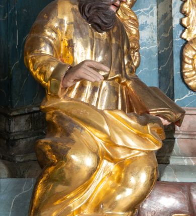 Zdjęcie nr 12: Ambona przyścienna, stojąca, z wejściem w postaci schodów z balustradą od strony prezbiterium. Kosz na planie ściętego z dwóch stron kwadratu, ustawiony pod kątem względem zaplecka, na profilowanym cokole, z podwieszonym u spodu zwężającym się w dół elementem podtrzymywanym przez figurę anioła. Dwie frontowe ścianki podzielone po środku pilastrami dźwigającymi pełne belkowanie o silnie wysuniętym gzymsie stanowiącym parapet kosza. Trzony pilastrów oraz pola pomiędzy nimi dekorowane są roślinnymi zwisami z muszlami. Na cokole kosza znajdują się siedzące na obłokach figury czterech ewangelistów z otwartymi księgami oraz na froncie figura Chrystusa trzymającego kulę. Podwieszony element pomiędzy siedzącymi figurami dekorowany jest obłokami i uskrzydlonymi główkami anielskimi. Balustrada schodów ozdobiona ornamentem roślinnym, zakończona półfilarem, na którym stoi figurka św. Jana Nepomucena. Zaplecek wąski, w formie stojącego prostokąta, z płyciną w polu ujętą profilowaną ramą w formie stojącego prostokąta zamkniętego łukiem segmentowym nadwieszonym z obrazem Chrystusa Dobrego Pasterza. Płycina flankowana rzędami kampanul, zwieńczona obłokiem z uskrzydloną główką anielską. Zaplecek z uszami z karbowanej wstęgi przeplatanej akantem, zwieńczony dwiema wolutami zwróconymi na zewnątrz. Całość zakończona ustawioną ukośnie glorią promienistą z gołębicą Ducha Świętego pośrodku. Struktura polichromowana na sposób imitujący błękitno-biało-różowy marmur. Dekoracja rzeźbiarska i ornamentalna pozłacana i posrebrzana, partie anatomiczne figur polichromowane naturalistycznie. 