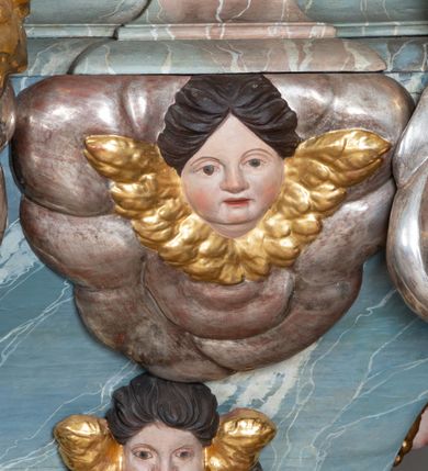 Zdjęcie nr 14: Ambona przyścienna, stojąca, z wejściem w postaci schodów z balustradą od strony prezbiterium. Kosz na planie ściętego z dwóch stron kwadratu, ustawiony pod kątem względem zaplecka, na profilowanym cokole, z podwieszonym u spodu zwężającym się w dół elementem podtrzymywanym przez figurę anioła. Dwie frontowe ścianki podzielone po środku pilastrami dźwigającymi pełne belkowanie o silnie wysuniętym gzymsie stanowiącym parapet kosza. Trzony pilastrów oraz pola pomiędzy nimi dekorowane są roślinnymi zwisami z muszlami. Na cokole kosza znajdują się siedzące na obłokach figury czterech ewangelistów z otwartymi księgami oraz na froncie figura Chrystusa trzymającego kulę. Podwieszony element pomiędzy siedzącymi figurami dekorowany jest obłokami i uskrzydlonymi główkami anielskimi. Balustrada schodów ozdobiona ornamentem roślinnym, zakończona półfilarem, na którym stoi figurka św. Jana Nepomucena. Zaplecek wąski, w formie stojącego prostokąta, z płyciną w polu ujętą profilowaną ramą w formie stojącego prostokąta zamkniętego łukiem segmentowym nadwieszonym z obrazem Chrystusa Dobrego Pasterza. Płycina flankowana rzędami kampanul, zwieńczona obłokiem z uskrzydloną główką anielską. Zaplecek z uszami z karbowanej wstęgi przeplatanej akantem, zwieńczony dwiema wolutami zwróconymi na zewnątrz. Całość zakończona ustawioną ukośnie glorią promienistą z gołębicą Ducha Świętego pośrodku. Struktura polichromowana na sposób imitujący błękitno-biało-różowy marmur. Dekoracja rzeźbiarska i ornamentalna pozłacana i posrebrzana, partie anatomiczne figur polichromowane naturalistycznie. 