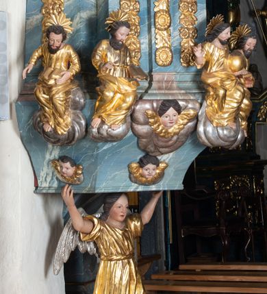 Zdjęcie nr 9: Ambona przyścienna, stojąca, z wejściem w postaci schodów z balustradą od strony prezbiterium. Kosz na planie ściętego z dwóch stron kwadratu, ustawiony pod kątem względem zaplecka, na profilowanym cokole, z podwieszonym u spodu zwężającym się w dół elementem podtrzymywanym przez figurę anioła. Dwie frontowe ścianki podzielone po środku pilastrami dźwigającymi pełne belkowanie o silnie wysuniętym gzymsie stanowiącym parapet kosza. Trzony pilastrów oraz pola pomiędzy nimi dekorowane są roślinnymi zwisami z muszlami. Na cokole kosza znajdują się siedzące na obłokach figury czterech ewangelistów z otwartymi księgami oraz na froncie figura Chrystusa trzymającego kulę. Podwieszony element pomiędzy siedzącymi figurami dekorowany jest obłokami i uskrzydlonymi główkami anielskimi. Balustrada schodów ozdobiona ornamentem roślinnym, zakończona półfilarem, na którym stoi figurka św. Jana Nepomucena. Zaplecek wąski, w formie stojącego prostokąta, z płyciną w polu ujętą profilowaną ramą w formie stojącego prostokąta zamkniętego łukiem segmentowym nadwieszonym z obrazem Chrystusa Dobrego Pasterza. Płycina flankowana rzędami kampanul, zwieńczona obłokiem z uskrzydloną główką anielską. Zaplecek z uszami z karbowanej wstęgi przeplatanej akantem, zwieńczony dwiema wolutami zwróconymi na zewnątrz. Całość zakończona ustawioną ukośnie glorią promienistą z gołębicą Ducha Świętego pośrodku. Struktura polichromowana na sposób imitujący błękitno-biało-różowy marmur. Dekoracja rzeźbiarska i ornamentalna pozłacana i posrebrzana, partie anatomiczne figur polichromowane naturalistycznie. 