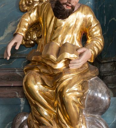 Zdjęcie nr 13: Ambona przyścienna, stojąca, z wejściem w postaci schodów z balustradą od strony prezbiterium. Kosz na planie ściętego z dwóch stron kwadratu, ustawiony pod kątem względem zaplecka, na profilowanym cokole, z podwieszonym u spodu zwężającym się w dół elementem podtrzymywanym przez figurę anioła. Dwie frontowe ścianki podzielone po środku pilastrami dźwigającymi pełne belkowanie o silnie wysuniętym gzymsie stanowiącym parapet kosza. Trzony pilastrów oraz pola pomiędzy nimi dekorowane są roślinnymi zwisami z muszlami. Na cokole kosza znajdują się siedzące na obłokach figury czterech ewangelistów z otwartymi księgami oraz na froncie figura Chrystusa trzymającego kulę. Podwieszony element pomiędzy siedzącymi figurami dekorowany jest obłokami i uskrzydlonymi główkami anielskimi. Balustrada schodów ozdobiona ornamentem roślinnym, zakończona półfilarem, na którym stoi figurka św. Jana Nepomucena. Zaplecek wąski, w formie stojącego prostokąta, z płyciną w polu ujętą profilowaną ramą w formie stojącego prostokąta zamkniętego łukiem segmentowym nadwieszonym z obrazem Chrystusa Dobrego Pasterza. Płycina flankowana rzędami kampanul, zwieńczona obłokiem z uskrzydloną główką anielską. Zaplecek z uszami z karbowanej wstęgi przeplatanej akantem, zwieńczony dwiema wolutami zwróconymi na zewnątrz. Całość zakończona ustawioną ukośnie glorią promienistą z gołębicą Ducha Świętego pośrodku. Struktura polichromowana na sposób imitujący błękitno-biało-różowy marmur. Dekoracja rzeźbiarska i ornamentalna pozłacana i posrebrzana, partie anatomiczne figur polichromowane naturalistycznie. 