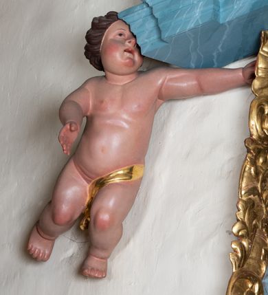 Zdjęcie nr 3: Ambona przyścienna, stojąca, z wejściem w postaci schodów z balustradą od strony prezbiterium. Kosz na planie ściętego z dwóch stron kwadratu, ustawiony pod kątem względem zaplecka, na profilowanym cokole, z podwieszonym u spodu zwężającym się w dół elementem podtrzymywanym przez figurę anioła. Dwie frontowe ścianki podzielone po środku pilastrami dźwigającymi pełne belkowanie o silnie wysuniętym gzymsie stanowiącym parapet kosza. Trzony pilastrów oraz pola pomiędzy nimi dekorowane są roślinnymi zwisami z muszlami. Na cokole kosza znajdują się siedzące na obłokach figury czterech ewangelistów z otwartymi księgami oraz na froncie figura Chrystusa trzymającego kulę. Podwieszony element pomiędzy siedzącymi figurami dekorowany jest obłokami i uskrzydlonymi główkami anielskimi. Balustrada schodów ozdobiona ornamentem roślinnym, zakończona półfilarem, na którym stoi figurka św. Jana Nepomucena. Zaplecek wąski, w formie stojącego prostokąta, z płyciną w polu ujętą profilowaną ramą w formie stojącego prostokąta zamkniętego łukiem segmentowym nadwieszonym z obrazem Chrystusa Dobrego Pasterza. Płycina flankowana rzędami kampanul, zwieńczona obłokiem z uskrzydloną główką anielską. Zaplecek z uszami z karbowanej wstęgi przeplatanej akantem, zwieńczony dwiema wolutami zwróconymi na zewnątrz. Całość zakończona ustawioną ukośnie glorią promienistą z gołębicą Ducha Świętego pośrodku. Struktura polichromowana na sposób imitujący błękitno-biało-różowy marmur. Dekoracja rzeźbiarska i ornamentalna pozłacana i posrebrzana, partie anatomiczne figur polichromowane naturalistycznie. 