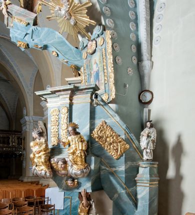 Zdjęcie nr 7: Ambona przyścienna, stojąca, z wejściem w postaci schodów z balustradą od strony prezbiterium. Kosz na planie ściętego z dwóch stron kwadratu, ustawiony pod kątem względem zaplecka, na profilowanym cokole, z podwieszonym u spodu zwężającym się w dół elementem podtrzymywanym przez figurę anioła. Dwie frontowe ścianki podzielone po środku pilastrami dźwigającymi pełne belkowanie o silnie wysuniętym gzymsie stanowiącym parapet kosza. Trzony pilastrów oraz pola pomiędzy nimi dekorowane są roślinnymi zwisami z muszlami. Na cokole kosza znajdują się siedzące na obłokach figury czterech ewangelistów z otwartymi księgami oraz na froncie figura Chrystusa trzymającego kulę. Podwieszony element pomiędzy siedzącymi figurami dekorowany jest obłokami i uskrzydlonymi główkami anielskimi. Balustrada schodów ozdobiona ornamentem roślinnym, zakończona półfilarem, na którym stoi figurka św. Jana Nepomucena. Zaplecek wąski, w formie stojącego prostokąta, z płyciną w polu ujętą profilowaną ramą w formie stojącego prostokąta zamkniętego łukiem segmentowym nadwieszonym z obrazem Chrystusa Dobrego Pasterza. Płycina flankowana rzędami kampanul, zwieńczona obłokiem z uskrzydloną główką anielską. Zaplecek z uszami z karbowanej wstęgi przeplatanej akantem, zwieńczony dwiema wolutami zwróconymi na zewnątrz. Całość zakończona ustawioną ukośnie glorią promienistą z gołębicą Ducha Świętego pośrodku. Struktura polichromowana na sposób imitujący błękitno-biało-różowy marmur. Dekoracja rzeźbiarska i ornamentalna pozłacana i posrebrzana, partie anatomiczne figur polichromowane naturalistycznie. 