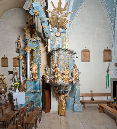 Zdjęcie nr 2: Ambona przyścienna, stojąca, z wejściem w postaci schodów z balustradą od strony prezbiterium. Kosz na planie ściętego z dwóch stron kwadratu, ustawiony pod kątem względem zaplecka, na profilowanym cokole, z podwieszonym u spodu zwężającym się w dół elementem podtrzymywanym przez figurę anioła. Dwie frontowe ścianki podzielone po środku pilastrami dźwigającymi pełne belkowanie o silnie wysuniętym gzymsie stanowiącym parapet kosza. Trzony pilastrów oraz pola pomiędzy nimi dekorowane są roślinnymi zwisami z muszlami. Na cokole kosza znajdują się siedzące na obłokach figury czterech ewangelistów z otwartymi księgami oraz na froncie figura Chrystusa trzymającego kulę. Podwieszony element pomiędzy siedzącymi figurami dekorowany jest obłokami i uskrzydlonymi główkami anielskimi. Balustrada schodów ozdobiona ornamentem roślinnym, zakończona półfilarem, na którym stoi figurka św. Jana Nepomucena. Zaplecek wąski, w formie stojącego prostokąta, z płyciną w polu ujętą profilowaną ramą w formie stojącego prostokąta zamkniętego łukiem segmentowym nadwieszonym z obrazem Chrystusa Dobrego Pasterza. Płycina flankowana rzędami kampanul, zwieńczona obłokiem z uskrzydloną główką anielską. Zaplecek z uszami z karbowanej wstęgi przeplatanej akantem, zwieńczony dwiema wolutami zwróconymi na zewnątrz. Całość zakończona ustawioną ukośnie glorią promienistą z gołębicą Ducha Świętego pośrodku. Struktura polichromowana na sposób imitujący błękitno-biało-różowy marmur. Dekoracja rzeźbiarska i ornamentalna pozłacana i posrebrzana, partie anatomiczne figur polichromowane naturalistycznie. 