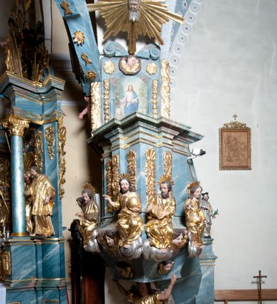 Zdjęcie nr 6: Ambona przyścienna, stojąca, z wejściem w postaci schodów z balustradą od strony prezbiterium. Kosz na planie ściętego z dwóch stron kwadratu, ustawiony pod kątem względem zaplecka, na profilowanym cokole, z podwieszonym u spodu zwężającym się w dół elementem podtrzymywanym przez figurę anioła. Dwie frontowe ścianki podzielone po środku pilastrami dźwigającymi pełne belkowanie o silnie wysuniętym gzymsie stanowiącym parapet kosza. Trzony pilastrów oraz pola pomiędzy nimi dekorowane są roślinnymi zwisami z muszlami. Na cokole kosza znajdują się siedzące na obłokach figury czterech ewangelistów z otwartymi księgami oraz na froncie figura Chrystusa trzymającego kulę. Podwieszony element pomiędzy siedzącymi figurami dekorowany jest obłokami i uskrzydlonymi główkami anielskimi. Balustrada schodów ozdobiona ornamentem roślinnym, zakończona półfilarem, na którym stoi figurka św. Jana Nepomucena. Zaplecek wąski, w formie stojącego prostokąta, z płyciną w polu ujętą profilowaną ramą w formie stojącego prostokąta zamkniętego łukiem segmentowym nadwieszonym z obrazem Chrystusa Dobrego Pasterza. Płycina flankowana rzędami kampanul, zwieńczona obłokiem z uskrzydloną główką anielską. Zaplecek z uszami z karbowanej wstęgi przeplatanej akantem, zwieńczony dwiema wolutami zwróconymi na zewnątrz. Całość zakończona ustawioną ukośnie glorią promienistą z gołębicą Ducha Świętego pośrodku. Struktura polichromowana na sposób imitujący błękitno-biało-różowy marmur. Dekoracja rzeźbiarska i ornamentalna pozłacana i posrebrzana, partie anatomiczne figur polichromowane naturalistycznie. 