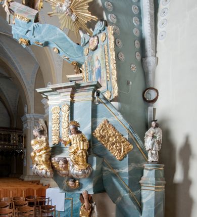 Zdjęcie nr 5: Ambona przyścienna, stojąca, z wejściem w postaci schodów z balustradą od strony prezbiterium. Kosz na planie ściętego z dwóch stron kwadratu, ustawiony pod kątem względem zaplecka, na profilowanym cokole, z podwieszonym u spodu zwężającym się w dół elementem podtrzymywanym przez figurę anioła. Dwie frontowe ścianki podzielone po środku pilastrami dźwigającymi pełne belkowanie o silnie wysuniętym gzymsie stanowiącym parapet kosza. Trzony pilastrów oraz pola pomiędzy nimi dekorowane są roślinnymi zwisami z muszlami. Na cokole kosza znajdują się siedzące na obłokach figury czterech ewangelistów z otwartymi księgami oraz na froncie figura Chrystusa trzymającego kulę. Podwieszony element pomiędzy siedzącymi figurami dekorowany jest obłokami i uskrzydlonymi główkami anielskimi. Balustrada schodów ozdobiona ornamentem roślinnym, zakończona półfilarem, na którym stoi figurka św. Jana Nepomucena. Zaplecek wąski, w formie stojącego prostokąta, z płyciną w polu ujętą profilowaną ramą w formie stojącego prostokąta zamkniętego łukiem segmentowym nadwieszonym z obrazem Chrystusa Dobrego Pasterza. Płycina flankowana rzędami kampanul, zwieńczona obłokiem z uskrzydloną główką anielską. Zaplecek z uszami z karbowanej wstęgi przeplatanej akantem, zwieńczony dwiema wolutami zwróconymi na zewnątrz. Całość zakończona ustawioną ukośnie glorią promienistą z gołębicą Ducha Świętego pośrodku. Struktura polichromowana na sposób imitujący błękitno-biało-różowy marmur. Dekoracja rzeźbiarska i ornamentalna pozłacana i posrebrzana, partie anatomiczne figur polichromowane naturalistycznie. 