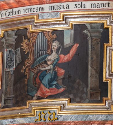 Zdjęcie nr 5: Balustrada chóru muzycznego o wklęsło-wypukłym zarysie, wybrzuszona w środkowej części, z cokołem i podwieszonym lambrekinem. Pola balustrady wydzielone pilastrami, o trzonach mających formę spływów wolutowych, dźwigającymi pełne, przełamane ponad nimi belkowanie, na którym, na osiach podpór, znajdują się kule. Cokół dekorowany rocaille'em i rozetami, a trzony pilastrów kampanulami. W polach znajdują się płyciny z malowanymi scenami. Scena środkowa przedstawia św. Cecylię grającą na organach we wnętrzu z oknem, ujętym filarami. Nad płyciną w partii fryzu belkowania i pod belkowaniem malowane banderole z napisami „Artes cum mundo cunctae pereunte peribunt,” i „In Coelum remeans musica sola manet.”, pod płyciną data „1771”. W czterech pionowych płycinach flankujących pole środkowe znajduje się płaskorzeźbiony rocaille. W pozostałych czterech poziomych płycinach malowane przedstawienia par muzykujących puttów stojących na rocaille'u. Polichromia balustrady imitująca marmur w kolorze szaro-niebieskim z marmoryzowanymi na czerwono profilami, ornamentyka i ramy płycin złocone i srebrzone.