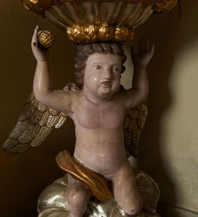 Zdjęcie nr 3: Na drewnianym cokole na rzucie koła znajduje się figura putta klęczącego na obłoku, podtrzymująca na głowie czarę chrzcielnicy z pokrywą. Putto ukazane jest frontalnie, klęczy na lewej nodze, prawą wyciąga do przodu, obie ręce ma uniesione w górę, w prawej dłoni trzyma owoc. Twarz ma kwadratową o pełnych policzkach, okoloną krótkimi włosami, skręconymi w pojedyncze pasma. W partii podbrzusza przesłonięte jest fragmentem tkaniny opadającej na prawe udo. Skrzydła ma złożone. Czara półkolista, imitująca puklowanie, zakończona gzymsem dekorowanym perełkowaniem, w połowie wysokości ujęta dekoracyjnym pasem z wolutowo skręconego akantu. Pokrywa na rzucie koła, dekorowana akantem, pełni funkcję podstawy, na której znajduje się niewielka rzeźbiarska grupa Chrztu Chrystusa z unoszącą się nad postaciami gołębicą Ducha Świętego w glorii promienistej. Ujęta jest sześcioma kabłąkami o wolutowych zakończeniach, podtrzymującymi cokół dla rzeźbionej półpostaci Boga Ojca ukazanego pośród obłoków, w glorii promienistej. Polichromia naturalistyczna w partiach ciała postaci, pozostałe elementy złocone i srebrzone. 