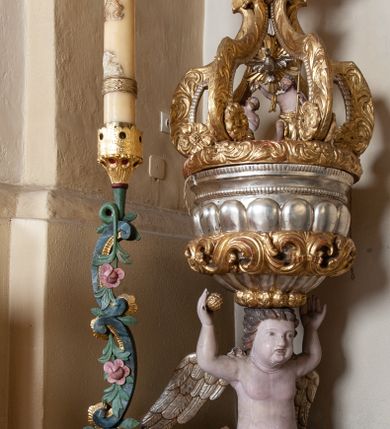 Zdjęcie nr 2: Na drewnianym cokole na rzucie koła znajduje się figura putta klęczącego na obłoku, podtrzymująca na głowie czarę chrzcielnicy z pokrywą. Putto ukazane jest frontalnie, klęczy na lewej nodze, prawą wyciąga do przodu, obie ręce ma uniesione w górę, w prawej dłoni trzyma owoc. Twarz ma kwadratową o pełnych policzkach, okoloną krótkimi włosami, skręconymi w pojedyncze pasma. W partii podbrzusza przesłonięte jest fragmentem tkaniny opadającej na prawe udo. Skrzydła ma złożone. Czara półkolista, imitująca puklowanie, zakończona gzymsem dekorowanym perełkowaniem, w połowie wysokości ujęta dekoracyjnym pasem z wolutowo skręconego akantu. Pokrywa na rzucie koła, dekorowana akantem, pełni funkcję podstawy, na której znajduje się niewielka rzeźbiarska grupa Chrztu Chrystusa z unoszącą się nad postaciami gołębicą Ducha Świętego w glorii promienistej. Ujęta jest sześcioma kabłąkami o wolutowych zakończeniach, podtrzymującymi cokół dla rzeźbionej półpostaci Boga Ojca ukazanego pośród obłoków, w glorii promienistej. Polichromia naturalistyczna w partiach ciała postaci, pozostałe elementy złocone i srebrzone. 