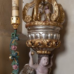 Zdjęcie nr 1: Na drewnianym cokole na rzucie koła znajduje się figura putta klęczącego na obłoku, podtrzymująca na głowie czarę chrzcielnicy z pokrywą. Putto ukazane jest frontalnie, klęczy na lewej nodze, prawą wyciąga do przodu, obie ręce ma uniesione w górę, w prawej dłoni trzyma owoc. Twarz ma kwadratową o pełnych policzkach, okoloną krótkimi włosami, skręconymi w pojedyncze pasma. W partii podbrzusza przesłonięte jest fragmentem tkaniny opadającej na prawe udo. Skrzydła ma złożone. Czara półkolista, imitująca puklowanie, zakończona gzymsem dekorowanym perełkowaniem, w połowie wysokości ujęta dekoracyjnym pasem z wolutowo skręconego akantu. Pokrywa na rzucie koła, dekorowana akantem, pełni funkcję podstawy, na której znajduje się niewielka rzeźbiarska grupa Chrztu Chrystusa z unoszącą się nad postaciami gołębicą Ducha Świętego w glorii promienistej. Ujęta jest sześcioma kabłąkami o wolutowych zakończeniach, podtrzymującymi cokół dla rzeźbionej półpostaci Boga Ojca ukazanego pośród obłoków, w glorii promienistej. Polichromia naturalistyczna w partiach ciała postaci, pozostałe elementy złocone i srebrzone. 