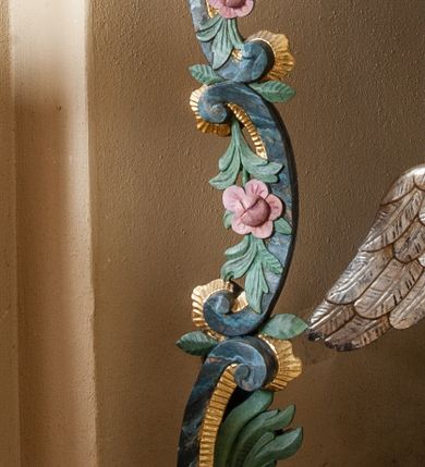 Zdjęcie nr 4: Na drewnianym cokole na rzucie koła znajduje się figura putta klęczącego na obłoku, podtrzymująca na głowie czarę chrzcielnicy z pokrywą. Putto ukazane jest frontalnie, klęczy na lewej nodze, prawą wyciąga do przodu, obie ręce ma uniesione w górę, w prawej dłoni trzyma owoc. Twarz ma kwadratową o pełnych policzkach, okoloną krótkimi włosami, skręconymi w pojedyncze pasma. W partii podbrzusza przesłonięte jest fragmentem tkaniny opadającej na prawe udo. Skrzydła ma złożone. Czara półkolista, imitująca puklowanie, zakończona gzymsem dekorowanym perełkowaniem, w połowie wysokości ujęta dekoracyjnym pasem z wolutowo skręconego akantu. Pokrywa na rzucie koła, dekorowana akantem, pełni funkcję podstawy, na której znajduje się niewielka rzeźbiarska grupa Chrztu Chrystusa z unoszącą się nad postaciami gołębicą Ducha Świętego w glorii promienistej. Ujęta jest sześcioma kabłąkami o wolutowych zakończeniach, podtrzymującymi cokół dla rzeźbionej półpostaci Boga Ojca ukazanego pośród obłoków, w glorii promienistej. Polichromia naturalistyczna w partiach ciała postaci, pozostałe elementy złocone i srebrzone. 