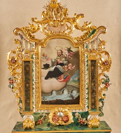 Zdjęcie nr 1: Podstawa na planie wydłużonego prostokąta, obwiedziona gzymsem, wyłamana w narożach, z otworami na drążki, dekorowana rozetami. Na niej zamocowana rama w formie zbliżonej do stojącego prostokąta, flankowana pilastrami ustawionymi na rocaille&#039;owych konsolach, o trzonach w formie przeszklonych gablot na relikwie na awersie oraz z rocaille&#039;m na rewersie oraz o rocaille&#039;owych kapitelach, dźwigających stylizowane imposty zakończone wolutami. Po bokach pilastrów rocaille z kwiatami. Rama zwieńczona szczytem utworzonym z rokokowego kartusza i anthemionu, ujętego rocaille&#039;m na awersie i rokokowego kartusza z nieczytelnym napisem „HR[...] / MI[...]” oraz niewielką półpostacią Boga Ojca na rewersie. U dołu, po obu stronach dekorowana rokokowym kartuszem flankowanym zwisami kwiatowymi. Pole obrazowe w formie stojącego prostokąta zamkniętego łukiem odcinkowym nadwieszonym, ujęte ramą dekorowaną motywem roślinnym. Na awersie obraz przedstawiający apoteozę św. Jana Nepomucena. W centrum postać świętego unoszącego się na obłokach, w pozycji siedzącej, zwróconego w trzech czwartych w lewo. Prawą rękę wyciąga przed siebie, lewą przytrzymuje na kolanach księgę i krucyfiks na ramieniu. Twarz ma owalną, o wysokim czole, dużych oczach skierowanych w górę, małym nosie i ustach, okoloną krótkimi, brązowymi włosami i brodą. Ubrany jest w czarną sutannę, białą komże lamowaną koronką oraz szarą almucję. Przed świętym na obłokach leży liść palmy i czarny biret, obok niego unosi się putto trzymając mu nad głową gwieździstą aureolę, pod nim putto trzyma kłódkę, a wokół niego unoszą się trzy uskrzydlone główki anielskie. Święty ukazany na tle obłoków. W dolnej części obrazu przedstawiony jest most Karola w Pradze na rzece Wełtawie, pośród pejzażu. Na rewersie płaskorzeźbione przedstawienie Chrystusa stojącego na globie dekorowanym kwiatami, ukazanego frontalnie, ze złożonymi z przodu dłońmi. Twarz ma pociągłą, o wysokim czole, dużych, migdałowatych oczach, dużym nosie i ustach, okoloną kręconymi włosami i brodą. Ubrany w srebrzoną tunikę oraz złocony płaszcz przewiązany przez prawe ramię i opadający po bokach postaci. Chrystusa w dolnej części przedstawienia flankują dwie rozety. Postać ukazana na laserowanym i cieniowanym, czerwonym tle. 





