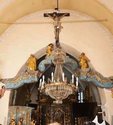 Zdjęcie nr 1: Belka tęczowa złożona z trzech elementów – dwóch bocznych w formie falistych i wyłamanych fragmentów belkowania, na których opiera się trzeci element w kształcie łuku kotarowego z uskokiem na ćwierćkolistych konsolach. Belka dekorowana rocaille&#039;m i rozetami, podtrzymywana przez dwie pary puttów.   Na zwieńczeniu łuku krzyż z figurą Chrystusa przymocowany do ściany tęczowej. Jezus ukazany jako umarły, przybity do krzyża trzema gwoździami. Jego ciało jest w lekkim zwisie, o szeroko rozłożonych ramionach, z głową opadająca bezwładnie na prawy bark, stopy skrzyżowane w układzie prawa na lewą. Chrystus ma podłużną twarz, o zamkniętych oczach, dużym nosie i szeroko otwartych ustach z widocznymi zębami; okoloną kręconymi i brązowymi włosami i brodą. Na głowie ma szeroką, ciemnozieloną koronę cierniową. Jego ciało jest silnie umięśnione, z podkreślonymi żebrami i mostkiem oraz zaznaczonymi śladami męki. Przez biodra ma przewiązane krótkie, srebrzone perizonium, silnie drapowane, ze zwisem tkaniny na lewym boku. Krzyż ciemnobrązowy, o prostym zakończeniu ramion, pionowa bela zwieńczona białą banderolą z napisem „INRI”. Pod krzyżem klęczy św. Maria Magdalena, zwrócona profilem w lewo, z pochyloną głową oraz wyciągniętymi do przodu rękami. Twarz ma owalna, o pełnych policzkach, małych oczach i nosie oraz otwartych ustach, okoloną długimi, falistymi włosami, opadającymi na plecy. Ubrana jest w złoconą suknię o szerokich, podwiniętych rękawach oraz złocony płaszcz zawinięty wokół pasa i nóg. Po prawej stronie krzyża stoi Matka Boska ukazana frontalnie, z uniesioną głową, lewą rękę kładzie na piersi, prawą wyciąga w bok. Twarz ma owalną, o oczach uniesionych w górę, małym nosie i ustach. Ubrana jest w złoconą suknię i płaszcz, na głowie ma złocony welon. Po lewej stronie krzyża stoi św. Jan Ewangelista, ukazany frontalnie, z uniesioną głową, prawą rękę kładzie na piersi, lewą wyciąga w bok. Twarz ma owalną, o oczach uniesionych w górę, małym nosie i ustach, okoloną krótkimi, brązowymi włosami. Ubrany jest w złoconą tunikę i płaszcz, zawinięty wokół bioder. Polichromia ciał postaci naturalistyczna. 



