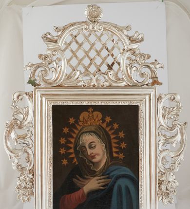 Zdjęcie nr 1: Rama srebrzona, w formie stojącego prostokąta, profilowana, dekorowana pasem ornamentu geometrycznego. Zwieńczona ażurowym szczytem utworzonym z fragmentu wyłamanego gzymsu, na konsolach, dekorowanego akantem i różami, wypełnionym kratką. Rama po bokach zdobiona akantem oraz kwiatowymi zwisami. Obraz przedstawia Matkę Boską w półpostaci, zwróconą w trzech czwartych w lewo, z pochyloną głową oraz skrzyżowanymi na piersi rękami. Twarz ma owalną, o dużych oczach skierowanych w dół, uniesionych brwiach, prostym nosie i pełnych ustach. Ubrana jest w czerwoną suknię, ciemnoniebieski płaszcz zarzucony na ramiona, wokół twarzy ma białą koronkę, a głowy brązowo-zielony welon, złoconą glorię oraz wieniec z gwiazd. Na głowie ma niewielką, złoconą koronę zamkniętą. Maria ukazana na jednolitym, brązowo-zielonym tle. 




