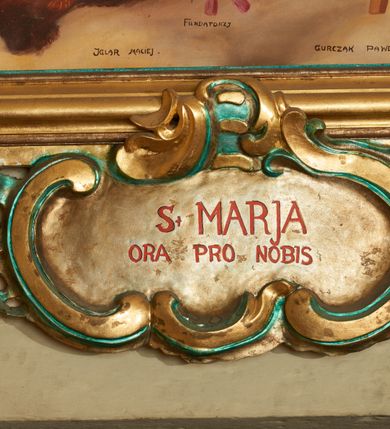 Zdjęcie nr 6: Obraz w formie stojącego prostokąta, ujęty profilowaną ramą, dekorowaną szczytem utworzonym z rocaille'owego kartusza z monogramem Marii w polu, flankowanym rocaille'm i kwiatami, podtrzymywanym w narożach konsolami. U dołu ramy podwieszenie utworzone z rocaille'owego kartusza z napisem „S(ANTA) MARIA / ORA PRO NOBIS”, ujęte rocaille'm i konsolami w narożach. Po bokach ramę trzymają nadlatujące putta. W centralnej części obrazu Matka Boska ukazana w pozycji stojącej pośród obłoków, z pochyloną głową i rozłożonymi na boki rękami podtrzymującymi płaszcz. Ubrana jest w różową suknię, przepasaną w talii, na ramionach ma obszerny, niebieski płaszcz, wokół głowy zawiązaną jasnobrązową chustę, na głowie koronę otwartą. W dolnej części kompozycji ukazane dwie grupy klęczących świętych, zwróconych do wewnątrz. Po lewej tronie św. Piotr. św. Stanisław i św. Stefan (?). Na pierwszym planie św. Stefan ukazany z profilu, z wyciągniętą w przód prawą ręką. Okolony jest krótką brodą i długimi, brązowymi włosami, ubrany w czerwony płaszcz królewski, ze złoconym wzorem, obszyty futrem. Przed nim na różowej poduszce z chwostami leży korona i jabłko królewskie. Obok niego św. Stanisław pozbawiony brody, w stroju biskupim, w żółtej kapie i infule oraz św. Piotr ukazany jako strzec, okolony siwymi włosami i brodą, w żółtym płaszczu. Po prawej stronie obrazu klęczą św. zakonnica, św. Katarzyna Aleksandryjska i św. Elżbieta Węgierska. Zakonnica ubrana jest w brązowy habit i welon oraz białą podwikę, prawą ręką przytrzymuje białą flagę, lewą wyciąga w bok. Obok niej klęczy św. Katarzyna z rekami złożonymi w geście modlitwy, o długich jasnych włosach. Ubrana jest w różowy płaszcz, na głowie ma koronę otwartą. Obok niej leży fragment koła. Za nimi postać św. Elżbiety, która podtrzymuje kwiaty róż. Ubrana jest w zielony płaszcz, na głowie ma czarny welon i koronę otwartą. Scena ukazana na tle jasnobłękitnego nieba wypełnionego żółto-brązowymi obłokami. W górnej części obrazu ukazane trzy putta. W dolnej części obrazu napis: „FUNDATORZY / MYSLIWIEC LUDWIK. IGLAR MACIEJ. GURCZYK PAWEŁ”. W prawym dolnym narożu obrazu sygnatura: „MAL(OWAŁ) KÛC ANTONI / 1932.”
