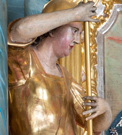Zdjęcie nr 4: Figura ścięta z tyłu, drążona, zwrócona w trzech czwartych w prawo, w kontrapoście. Święty obiema rękami trzyma włócznię skierowaną w dół w stronę leżącego u jego stóp diabła. Twarz ma pociągłą, o migdałowatych oczach, lekko garbatym nosie, pełnych ustach, okoloną długimi, silnie kręconymi włosami. Postać w stroju żołnierza, w tunice sięgającej kolan, zbroi oraz w płaszczu zarzuconym na plecy i opadającym na prawą nogę, przewiązanym taśmą przez tors, na głowie ma hełm. Ciało polichromowane naturalistycznie, strój złocony. 
