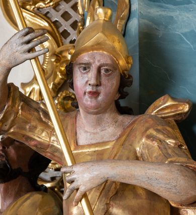 Zdjęcie nr 3: Figura ścięta z tyłu, drążona, zwrócona w trzech czwartych w prawo, w kontrapoście. Święty obiema rękami trzyma włócznię skierowaną w dół w stronę leżącego u jego stóp diabła. Twarz ma pociągłą, o migdałowatych oczach, lekko garbatym nosie, pełnych ustach, okoloną długimi, silnie kręconymi włosami. Postać w stroju żołnierza, w tunice sięgającej kolan, zbroi oraz w płaszczu zarzuconym na plecy i opadającym na prawą nogę, przewiązanym taśmą przez tors, na głowie ma hełm. Ciało polichromowane naturalistycznie, strój złocony. 

