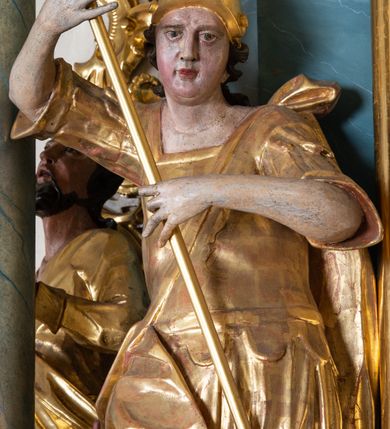 Zdjęcie nr 2: Figura ścięta z tyłu, drążona, zwrócona w trzech czwartych w prawo, w kontrapoście. Święty obiema rękami trzyma włócznię skierowaną w dół w stronę leżącego u jego stóp diabła. Twarz ma pociągłą, o migdałowatych oczach, lekko garbatym nosie, pełnych ustach, okoloną długimi, silnie kręconymi włosami. Postać w stroju żołnierza, w tunice sięgającej kolan, zbroi oraz w płaszczu zarzuconym na plecy i opadającym na prawą nogę, przewiązanym taśmą przez tors, na głowie ma hełm. Ciało polichromowane naturalistycznie, strój złocony. 
