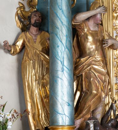 Zdjęcie nr 5: Figura ścięta z tyłu, drążona, zwrócona w trzech czwartych w prawo, w kontrapoście. Święty obiema rękami trzyma włócznię skierowaną w dół w stronę leżącego u jego stóp diabła. Twarz ma pociągłą, o migdałowatych oczach, lekko garbatym nosie, pełnych ustach, okoloną długimi, silnie kręconymi włosami. Postać w stroju żołnierza, w tunice sięgającej kolan, zbroi oraz w płaszczu zarzuconym na plecy i opadającym na prawą nogę, przewiązanym taśmą przez tors, na głowie ma hełm. Ciało polichromowane naturalistycznie, strój złocony. 
