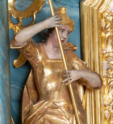 Zdjęcie nr 1: Figura ścięta z tyłu, drążona, zwrócona w trzech czwartych w prawo, w kontrapoście. Święty obiema rękami trzyma włócznię skierowaną w dół w stronę leżącego u jego stóp diabła. Twarz ma pociągłą, o migdałowatych oczach, lekko garbatym nosie, pełnych ustach, okoloną długimi, silnie kręconymi włosami. Postać w stroju żołnierza, w tunice sięgającej kolan, zbroi oraz w płaszczu zarzuconym na plecy i opadającym na prawą nogę, przewiązanym taśmą przez tors, na głowie ma hełm. Ciało polichromowane naturalistycznie, strój złocony. 
