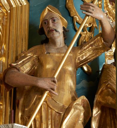 Zdjęcie nr 1: Figura ścięta z tyłu, drążona, zwrócona frontalnie, z głową lekko skierowaną w lewo, w kontrapoście. Święty obiema rękami trzyma włócznię skierowaną w stronę smoka, leżącego u jego stóp. Twarz ma pociągłą, o zapadniętych policzkach, oczach migdałowatych, nosie lekko garbatym, ustach pełnych, okoloną wąskimi, długimi wąsami, włosy długie, silnie kręcone, opadające na plecy. Ubrany jest w strój żołnierza: tunikę sięgającą kolan, rzymską zbroję z piórami oraz płaszcz narzucony na plecy i opadający na lewą nogę, na głowie ma hełm. Ciało polichromowane naturalistycznie, strój złocony.
