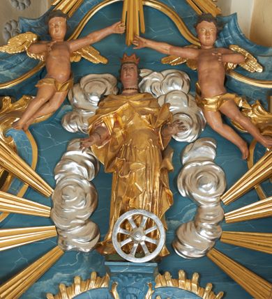 Zdjęcie nr 1: Figura ścięta z tyłu. Święta przedstawiona jest w całej postaci, stoi frontalnie, w delikatnym kontrapoście, z wysuniętym w bok lewym biodrem. Obie ręce ma ugięte i wyciągnięte w przód. Jej twarz jest pociągła o pełnych policzkach, z uwypukloną brodą i podbródkiem oraz niskim czołem. Oczy ma migdałowate, nos prosty, usta pełne, lekko rozchylone. Jej twarz jest okolona długimi, mocno falowanymi włosami opadającymi na plecy. Święta ubrana jest w suknię z dekoltem, spiętą gorsetem w talii oraz płaszcz zapięty na piersi i opadający na boki postaci, z fragmentem tkaniny zawiniętym na jej prawej nodze. Na głowie ma koronę otwartą, na szyi i nadgarstku biżuterię. Przed figurą znajduje się koło. Polichromia naturalistyczna w partiach ciała, strój złocony, koło srebrzone.