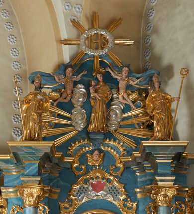 Zdjęcie nr 2: Figura ścięta z tyłu. Święta przedstawiona jest w całej postaci, stoi frontalnie, w delikatnym kontrapoście, z wysuniętym w bok lewym biodrem. Obie ręce ma ugięte i wyciągnięte w przód. Jej twarz jest pociągła o pełnych policzkach, z uwypukloną brodą i podbródkiem oraz niskim czołem. Oczy ma migdałowate, nos prosty, usta pełne, lekko rozchylone. Jej twarz jest okolona długimi, mocno falowanymi włosami opadającymi na plecy. Święta ubrana jest w suknię z dekoltem, spiętą gorsetem w talii oraz płaszcz zapięty na piersi i opadający na boki postaci, z fragmentem tkaniny zawiniętym na jej prawej nodze. Na głowie ma koronę otwartą, na szyi i nadgarstku biżuterię. Przed figurą znajduje się koło. Polichromia naturalistyczna w partiach ciała, strój złocony, koło srebrzone.