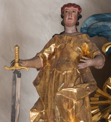 Zdjęcie nr 1: Figura ścięta z tyłu. Święta przedstawiona jest w całej postaci, stojąca frontalnie, z lekko ugiętą lewą nogą. W prawej dłoni trzyma miecz zwrócony ostrzem w dół, lewą rękę wyciąga przed siebie. Twarz ma podłużną o pełnych policzkach i drugim podbródku, migdałowatych oczach, prostym nosie, okoloną silnie kręconymi włosami opadającymi na plecy. Święta ubrana jest w suknię z dekoltem, z podwiniętymi rękawami, spiętą w talii gorsetem oraz płaszcz opadający na plecy i lewy bok postaci. Na głowie ma wieniec z róż. Polichromia naturalistyczna w partiach ciała, szaty złocone, ostrze miecza srebrzone. 

