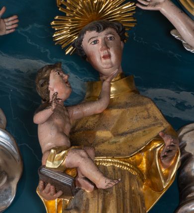 Zdjęcie nr 1: Figura ścięta z tyłu. Święty przedstawiony jest w całej postaci, frontalnie, w kontrapoście, delikatnie przechylony w tył. Lewą rękę zgiętą w łokciu wyciąga w przód, w prawej, zgiętej trzyma księgę, na której siedzi Dzieciątko. Twarz ma pociągłą, o pełnych policzkach, migdałowatych oczach, prostym nosie i pełnych ustach, z silnie zaznaczoną brodą i podbródkiem, okoloną krótkimi, kręconymi włosami. Święty ubrany jest w habit przewiązany w pasie podwójnym sznurem i pelerynkę. Nad jego głową nimb w formie glorii promienistej. Dzieciątko zwrócone jest profilem w prawo, lewą ręką ujmuje twarz świętego, prawą wyciąga w bok. Twarz ma pociągłą o pełnych policzkach, okoloną kręconymi włosami, ciało zaś ma szczupłe, w partii bioder przykryte tkaniną. Polichromia naturalistyczna w partiach ciała, szaty i gloria pozłocone.
