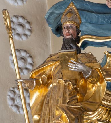 Zdjęcie nr 1: Figura ścięta z tyłu. Święty przedstawiony jest w całej postaci, frontalnie, w kontrapoście. W swojej lewej, uniesionej ręce trzyma zamkniętą księgę, zaś w prawej dłoni, wyciągniętej w bok, pastorał. Twarz ma pociągłą, o dużych oczach, prostym nosie i otwartych ustach, okoloną długą brodą z wąskimi wąsami oraz silnie kręconymi krótkimi włosami. Święty ubrany jest w długą albę, przepasaną w talii oraz kapę zapiętą na piersi, okrywającą ramiona z rozwianą fałdą zawiniętą na jego lewej nodze, na głowie ma infułę, a na dłoniach rękawiczki. Polichromia naturalistyczna w partiach ciała, szaty i atrybuty pozłocone, detale stroju posrebrzone. 