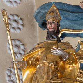 Zdjęcie nr 1: Figura ścięta z tyłu. Święty przedstawiony jest w całej postaci, frontalnie, w kontrapoście. W swojej lewej, uniesionej ręce trzyma zamkniętą księgę, zaś w prawej dłoni, wyciągniętej w bok, pastorał. Twarz ma pociągłą, o dużych oczach, prostym nosie i otwartych ustach, okoloną długą brodą z wąskimi wąsami oraz silnie kręconymi krótkimi włosami. Święty ubrany jest w długą albę, przepasaną w talii oraz kapę zapiętą na piersi, okrywającą ramiona z rozwianą fałdą zawiniętą na jego lewej nodze, na głowie ma infułę, a na dłoniach rękawiczki. Polichromia naturalistyczna w partiach ciała, szaty i atrybuty pozłocone, detale stroju posrebrzone. 