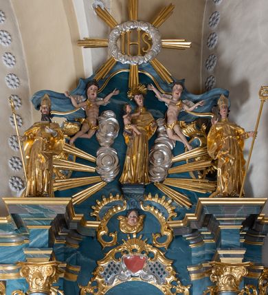 Zdjęcie nr 2: Figura ścięta z tyłu. Święty przedstawiony jest w całej postaci, frontalnie, w kontrapoście. W swojej lewej, uniesionej ręce trzyma zamkniętą księgę, zaś w prawej dłoni, wyciągniętej w bok, pastorał. Twarz ma pociągłą, o dużych oczach, prostym nosie i otwartych ustach, okoloną długą brodą z wąskimi wąsami oraz silnie kręconymi krótkimi włosami. Święty ubrany jest w długą albę, przepasaną w talii oraz kapę zapiętą na piersi, okrywającą ramiona z rozwianą fałdą zawiniętą na jego lewej nodze, na głowie ma infułę, a na dłoniach rękawiczki. Polichromia naturalistyczna w partiach ciała, szaty i atrybuty pozłocone, detale stroju posrebrzone. 