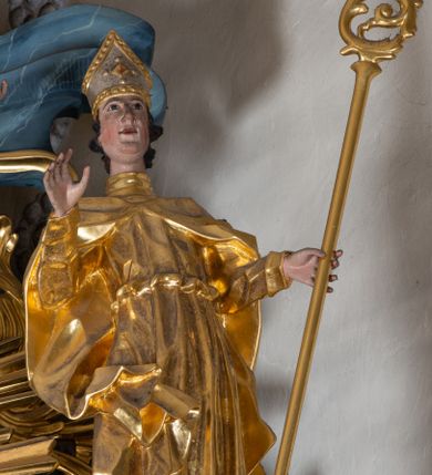 Zdjęcie nr 1: Figura ścięta z tyłu. Święty przedstawiony jest w całej postaci, frontalnie, w kontrapoście, jego sylwetka jest esowato wygięta. Prawa ręka jest uniesiona, w lewej, wyciągniętej w bok trzyma pastorał. Twarz ma podłużną, o migdałowatych oczach, prostym nosie, rozchylonych ustach i podkreślonej, wystającej brodzie, okoloną krótkimi, falistymi włosami. Święty ubrany jest w długą albę, przepasaną w talii oraz kapę spiętą pod szyją, okrywającą ramiona z rozwianą fałdą zawiniętą przy jego prawej nodze, na głowie ma infułę. Polichromia naturalistyczna w partiach ciała, szaty i atrybuty złocone, detale stroju srebrzone.