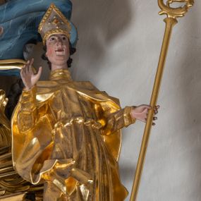 Zdjęcie nr 1: Figura ścięta z tyłu. Święty przedstawiony jest w całej postaci, frontalnie, w kontrapoście, jego sylwetka jest esowato wygięta. Prawa ręka jest uniesiona, w lewej, wyciągniętej w bok trzyma pastorał. Twarz ma podłużną, o migdałowatych oczach, prostym nosie, rozchylonych ustach i podkreślonej, wystającej brodzie, okoloną krótkimi, falistymi włosami. Święty ubrany jest w długą albę, przepasaną w talii oraz kapę spiętą pod szyją, okrywającą ramiona z rozwianą fałdą zawiniętą przy jego prawej nodze, na głowie ma infułę. Polichromia naturalistyczna w partiach ciała, szaty i atrybuty złocone, detale stroju srebrzone.