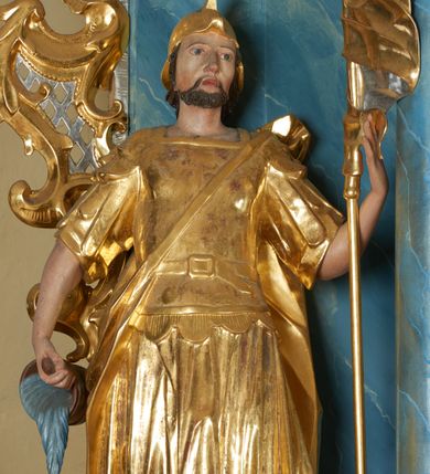 Zdjęcie nr 2: Rzeźba ścięta z tyłu, drążona, zwrócona frontalnie, w wykroku. Święty w lewej ręce trzyma chorągiew zakończoną ostrzem włóczni, w prawej naczynie z wodą, którą polewa niewielki, palący się dom znajdujący się przy jego prawej nodze. Twarz ma pociągłą, o zapadniętych policzkach, migdałowatych oczach, lekko garbatym nosie i pełnych ustach, okoloną wąsami, krótką brodą i włosami do ramion. Ubrany jest w strój rzymskiego żołnierza: tunikę sięgającą kolan, rzymską zbroję z piórami, płaszcz narzucony na plecy, podtrzymywany na taśmie przeciągniętej przez tors, na głowie nosi hełm, na nogach wysokie sandały. Ciało polichromowane naturalistycznie, szaty złocone. 