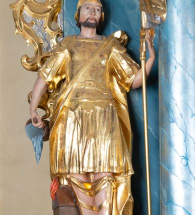 Zdjęcie nr 1: Rzeźba ścięta z tyłu, drążona, zwrócona frontalnie, w wykroku. Święty w lewej ręce trzyma chorągiew zakończoną ostrzem włóczni, w prawej naczynie z wodą, którą polewa niewielki, palący się dom znajdujący się przy jego prawej nodze. Twarz ma pociągłą, o zapadniętych policzkach, migdałowatych oczach, lekko garbatym nosie i pełnych ustach, okoloną wąsami, krótką brodą i włosami do ramion. Ubrany jest w strój rzymskiego żołnierza: tunikę sięgającą kolan, rzymską zbroję z piórami, płaszcz narzucony na plecy, podtrzymywany na taśmie przeciągniętej przez tors, na głowie nosi hełm, na nogach wysokie sandały. Ciało polichromowane naturalistycznie, szaty złocone. 