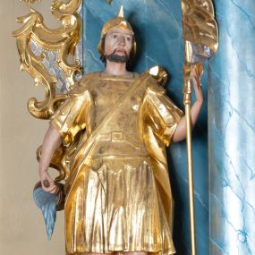 Zdjęcie nr 1: Rzeźba ścięta z tyłu, drążona, zwrócona frontalnie, w wykroku. Święty w lewej ręce trzyma chorągiew zakończoną ostrzem włóczni, w prawej naczynie z wodą, którą polewa niewielki, palący się dom znajdujący się przy jego prawej nodze. Twarz ma pociągłą, o zapadniętych policzkach, migdałowatych oczach, lekko garbatym nosie i pełnych ustach, okoloną wąsami, krótką brodą i włosami do ramion. Ubrany jest w strój rzymskiego żołnierza: tunikę sięgającą kolan, rzymską zbroję z piórami, płaszcz narzucony na plecy, podtrzymywany na taśmie przeciągniętej przez tors, na głowie nosi hełm, na nogach wysokie sandały. Ciało polichromowane naturalistycznie, szaty złocone. 