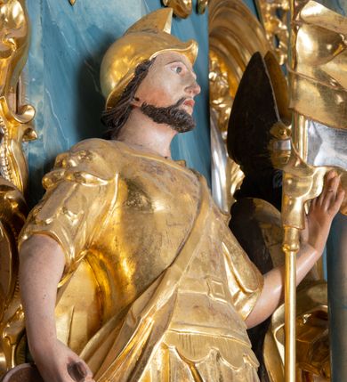 Zdjęcie nr 3: Rzeźba ścięta z tyłu, drążona, zwrócona frontalnie, w wykroku. Święty w lewej ręce trzyma chorągiew zakończoną ostrzem włóczni, w prawej naczynie z wodą, którą polewa niewielki, palący się dom znajdujący się przy jego prawej nodze. Twarz ma pociągłą, o zapadniętych policzkach, migdałowatych oczach, lekko garbatym nosie i pełnych ustach, okoloną wąsami, krótką brodą i włosami do ramion. Ubrany jest w strój rzymskiego żołnierza: tunikę sięgającą kolan, rzymską zbroję z piórami, płaszcz narzucony na plecy, podtrzymywany na taśmie przeciągniętej przez tors, na głowie nosi hełm, na nogach wysokie sandały. Ciało polichromowane naturalistycznie, szaty złocone. 