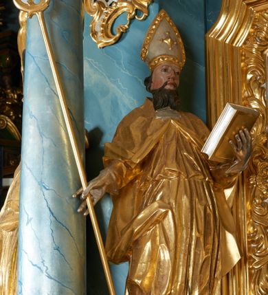 Zdjęcie nr 1: Rzeźba ścięta z tyłu, drążona, zwrócona frontalnie, w kontrapoście, lekko przechylona w tył. Święty w lewej ręce podtrzymuje zamkniętą księgę, w prawej trzyma pastorał. Twarz ma pociągłą o migdałowatych oczach, lekko garbatym nosie, pełnych ustach, okoloną wąsami i falistą brodą oraz krótkimi, kręconymi włosami. Postać ubrana jest w albę przewiązaną w pasie oraz kapę zapiętą na piersi, z połą tkaniny zawiniętą na prawej nodze, na głowie ma infułę, na dłoniach rękawiczki. Ciało polichromowane naturalistycznie, szaty oraz atrybuty złocone i srebrzone. 