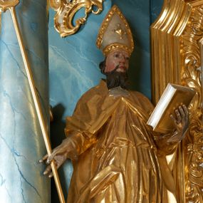 Zdjęcie nr 1: Rzeźba ścięta z tyłu, drążona, zwrócona frontalnie, w kontrapoście, lekko przechylona w tył. Święty w lewej ręce podtrzymuje zamkniętą księgę, w prawej trzyma pastorał. Twarz ma pociągłą o migdałowatych oczach, lekko garbatym nosie, pełnych ustach, okoloną wąsami i falistą brodą oraz krótkimi, kręconymi włosami. Postać ubrana jest w albę przewiązaną w pasie oraz kapę zapiętą na piersi, z połą tkaniny zawiniętą na prawej nodze, na głowie ma infułę, na dłoniach rękawiczki. Ciało polichromowane naturalistycznie, szaty oraz atrybuty złocone i srebrzone. 