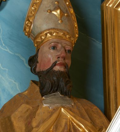 Zdjęcie nr 2: Rzeźba ścięta z tyłu, drążona, zwrócona frontalnie, w kontrapoście, lekko przechylona w tył. Święty w lewej ręce podtrzymuje zamkniętą księgę, w prawej trzyma pastorał. Twarz ma pociągłą o migdałowatych oczach, lekko garbatym nosie, pełnych ustach, okoloną wąsami i falistą brodą oraz krótkimi, kręconymi włosami. Postać ubrana jest w albę przewiązaną w pasie oraz kapę zapiętą na piersi, z połą tkaniny zawiniętą na prawej nodze, na głowie ma infułę, na dłoniach rękawiczki. Ciało polichromowane naturalistycznie, szaty oraz atrybuty złocone i srebrzone. 