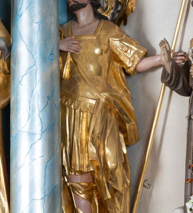 Zdjęcie nr 2: Rzeźba ścięta z tyłu, drążona, zwrócona frontalnie, z głową skierowaną w prawo, w kontrapoście. Święty w lewej ręce trzyma włócznię, prawa spoczywa na piersi. Twarz ma okrągłą o migdałowatych oczach, lekko garbatym nosie i pełnych ustach, okoloną krótką brodą i wąsami oraz długimi silnie skręconymi włosami opadającymi na plecy. Postać ubrana jest w tunikę sięgającą kolan, rzymską zbroję z piórami oraz płaszcz zawieszony na taśmie, opadający na lewy bok. Na głowie ma wieniec laurowy, na nogach wysokie sandały. Ciało polichromowane naturalistycznie, szaty i atrybuty złocone. 