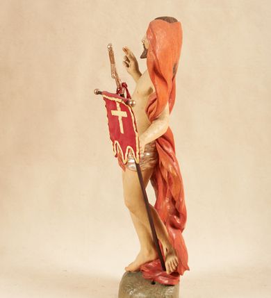 Zdjęcie nr 8: Rzeźba pełnoplastyczna, ustawiona na podstawie o nieregularnym planie, imitującej fragment podłoża. Chrystus ukazany frontalnie, w wykroku, w lewej wyciągniętej w bok ręce trzyma drzewce czerwonej, materiałowej chorągwi, zakończone krzyżem, prawą ugiętą, unosi w geście błogosławieństwa. Twarz ma szczupłą i pociągłą, o wąskich, migdałowatych oczach, szerokim nosie i niewielkich ustach; okoloną krótką brodą oraz silnie skręconymi w pukle włosami, opadającymi na ramiona. Chrystus w partii bioder przewiązany krótkim, przylegającym, srebrzonym perizonium. Przez prawe udo i lewe ramię ma przewiązany czerwony, silnie drapowany płaszcz, rozwiany półkoliście z tyłu postaci oraz opadający po bokach. Ciało Chrystusa szczupłe, niemal pozbawione modelunku. Polichromia ciała figury naturalistyczna, o jasnej karnacji. 



