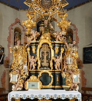 Zdjęcie nr 10: Ołtarz dwustronny, sarkofagowy, dekorowany rocaille'm, mensa drewniana na planie kwadratu, od frontu z kamiennym portatylem.
Nastawa ołtarzowa dwustronna, na rzucie leżącej litery X, w formie  rozbudowanego tabernakulum z tronem wystawienia. Obie strony są analogiczne pod względem architektonicznym. Tabernakulum na cokole, o wypukłej ściance frontowej, flankowanej parą pilastrów o trzonach dekorowanym płycinami z rocaille'm, dźwigających gzyms wieńczący. W polu drzwiczki ujęte rokokowym kartuszem. Na tabernakulum tron wystawienia o wklęsło-wypukłym licu, ujęty parą impostów podtrzymywanych przez rocaille'owe konsole oraz pilastrami w narożach, których trzony przyjmują formę spływów wolutowych, sięgające podstawy tabernakulum, na których stoją figury. W polu środkowym tronu wystawienia znajduje się obrotowa nisza o nieregularnym kształcie ujęta obramieniem z rocaille'u, przesłonięta wypukłą ścianką z płaskorzeźbionym krucyfiksem  i winnym gronem, zwieńczona rzeźbioną półpostacią. Całość wieńczy cokół z dwoma ramionami, na których stoją rocaille'owe wazony. Na cokole osadzona jest dużych rozmiarów, dwustronna, dwunastoramienna gwiazda relikwiarzowa, miażdżąca węża. Gwiazda z obu stron ujęta jest kilkoma pierścieniami z winnego grona oraz ornamentu roślinnego, w ramionach oraz w polu głównym umieszczone są za szkłem relikwie wśród kwiatowych dekoracji. 
Na awersie ołtarza, na mensie znajdują się dwie figurki puttów siedzących na obłokach i trzymających tabliczki z kanonami. Całość flankują ustawione na spływach wolutowych figury św. Jana Ewangelisty i św. Bartłomieja, tron wieńczy półpostać Boga Ojca, ujęta przez dwa putta z otwartymi księgami. Obrotową niszę flankują dwa aniołki trzymające lichtarze. Na rewersie ołtarza na spływach wolutowych stoją figury św. Andrzeja i św. Piotra, tron wieńczy półpostać Mojżesza flankowana przez dwa putta z otwartymi księgami.
Struktura polichromowana w kolorze ciemnozielonym, ornamentyka i profile złocone, detal srebrzony.
