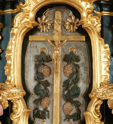 Zdjęcie nr 15: Ołtarz dwustronny, sarkofagowy, dekorowany rocaille'm, mensa drewniana na planie kwadratu, od frontu z kamiennym portatylem.
Nastawa ołtarzowa dwustronna, na rzucie leżącej litery X, w formie  rozbudowanego tabernakulum z tronem wystawienia. Obie strony są analogiczne pod względem architektonicznym. Tabernakulum na cokole, o wypukłej ściance frontowej, flankowanej parą pilastrów o trzonach dekorowanym płycinami z rocaille'm, dźwigających gzyms wieńczący. W polu drzwiczki ujęte rokokowym kartuszem. Na tabernakulum tron wystawienia o wklęsło-wypukłym licu, ujęty parą impostów podtrzymywanych przez rocaille'owe konsole oraz pilastrami w narożach, których trzony przyjmują formę spływów wolutowych, sięgające podstawy tabernakulum, na których stoją figury. W polu środkowym tronu wystawienia znajduje się obrotowa nisza o nieregularnym kształcie ujęta obramieniem z rocaille'u, przesłonięta wypukłą ścianką z płaskorzeźbionym krucyfiksem  i winnym gronem, zwieńczona rzeźbioną półpostacią. Całość wieńczy cokół z dwoma ramionami, na których stoją rocaille'owe wazony. Na cokole osadzona jest dużych rozmiarów, dwustronna, dwunastoramienna gwiazda relikwiarzowa, miażdżąca węża. Gwiazda z obu stron ujęta jest kilkoma pierścieniami z winnego grona oraz ornamentu roślinnego, w ramionach oraz w polu głównym umieszczone są za szkłem relikwie wśród kwiatowych dekoracji. 
Na awersie ołtarza, na mensie znajdują się dwie figurki puttów siedzących na obłokach i trzymających tabliczki z kanonami. Całość flankują ustawione na spływach wolutowych figury św. Jana Ewangelisty i św. Bartłomieja, tron wieńczy półpostać Boga Ojca, ujęta przez dwa putta z otwartymi księgami. Obrotową niszę flankują dwa aniołki trzymające lichtarze. Na rewersie ołtarza na spływach wolutowych stoją figury św. Andrzeja i św. Piotra, tron wieńczy półpostać Mojżesza flankowana przez dwa putta z otwartymi księgami.
Struktura polichromowana w kolorze ciemnozielonym, ornamentyka i profile złocone, detal srebrzony.
