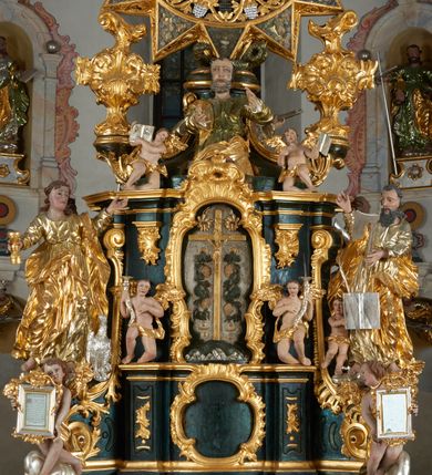 Zdjęcie nr 13: Ołtarz dwustronny, sarkofagowy, dekorowany rocaille'm, mensa drewniana na planie kwadratu, od frontu z kamiennym portatylem.
Nastawa ołtarzowa dwustronna, na rzucie leżącej litery X, w formie  rozbudowanego tabernakulum z tronem wystawienia. Obie strony są analogiczne pod względem architektonicznym. Tabernakulum na cokole, o wypukłej ściance frontowej, flankowanej parą pilastrów o trzonach dekorowanym płycinami z rocaille'm, dźwigających gzyms wieńczący. W polu drzwiczki ujęte rokokowym kartuszem. Na tabernakulum tron wystawienia o wklęsło-wypukłym licu, ujęty parą impostów podtrzymywanych przez rocaille'owe konsole oraz pilastrami w narożach, których trzony przyjmują formę spływów wolutowych, sięgające podstawy tabernakulum, na których stoją figury. W polu środkowym tronu wystawienia znajduje się obrotowa nisza o nieregularnym kształcie ujęta obramieniem z rocaille'u, przesłonięta wypukłą ścianką z płaskorzeźbionym krucyfiksem  i winnym gronem, zwieńczona rzeźbioną półpostacią. Całość wieńczy cokół z dwoma ramionami, na których stoją rocaille'owe wazony. Na cokole osadzona jest dużych rozmiarów, dwustronna, dwunastoramienna gwiazda relikwiarzowa, miażdżąca węża. Gwiazda z obu stron ujęta jest kilkoma pierścieniami z winnego grona oraz ornamentu roślinnego, w ramionach oraz w polu głównym umieszczone są za szkłem relikwie wśród kwiatowych dekoracji. 
Na awersie ołtarza, na mensie znajdują się dwie figurki puttów siedzących na obłokach i trzymających tabliczki z kanonami. Całość flankują ustawione na spływach wolutowych figury św. Jana Ewangelisty i św. Bartłomieja, tron wieńczy półpostać Boga Ojca, ujęta przez dwa putta z otwartymi księgami. Obrotową niszę flankują dwa aniołki trzymające lichtarze. Na rewersie ołtarza na spływach wolutowych stoją figury św. Andrzeja i św. Piotra, tron wieńczy półpostać Mojżesza flankowana przez dwa putta z otwartymi księgami.
Struktura polichromowana w kolorze ciemnozielonym, ornamentyka i profile złocone, detal srebrzony.
