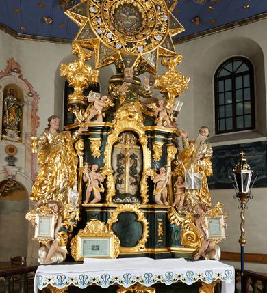 Zdjęcie nr 11: Ołtarz dwustronny, sarkofagowy, dekorowany rocaille'm, mensa drewniana na planie kwadratu, od frontu z kamiennym portatylem.
Nastawa ołtarzowa dwustronna, na rzucie leżącej litery X, w formie  rozbudowanego tabernakulum z tronem wystawienia. Obie strony są analogiczne pod względem architektonicznym. Tabernakulum na cokole, o wypukłej ściance frontowej, flankowanej parą pilastrów o trzonach dekorowanym płycinami z rocaille'm, dźwigających gzyms wieńczący. W polu drzwiczki ujęte rokokowym kartuszem. Na tabernakulum tron wystawienia o wklęsło-wypukłym licu, ujęty parą impostów podtrzymywanych przez rocaille'owe konsole oraz pilastrami w narożach, których trzony przyjmują formę spływów wolutowych, sięgające podstawy tabernakulum, na których stoją figury. W polu środkowym tronu wystawienia znajduje się obrotowa nisza o nieregularnym kształcie ujęta obramieniem z rocaille'u, przesłonięta wypukłą ścianką z płaskorzeźbionym krucyfiksem  i winnym gronem, zwieńczona rzeźbioną półpostacią. Całość wieńczy cokół z dwoma ramionami, na których stoją rocaille'owe wazony. Na cokole osadzona jest dużych rozmiarów, dwustronna, dwunastoramienna gwiazda relikwiarzowa, miażdżąca węża. Gwiazda z obu stron ujęta jest kilkoma pierścieniami z winnego grona oraz ornamentu roślinnego, w ramionach oraz w polu głównym umieszczone są za szkłem relikwie wśród kwiatowych dekoracji. 
Na awersie ołtarza, na mensie znajdują się dwie figurki puttów siedzących na obłokach i trzymających tabliczki z kanonami. Całość flankują ustawione na spływach wolutowych figury św. Jana Ewangelisty i św. Bartłomieja, tron wieńczy półpostać Boga Ojca, ujęta przez dwa putta z otwartymi księgami. Obrotową niszę flankują dwa aniołki trzymające lichtarze. Na rewersie ołtarza na spływach wolutowych stoją figury św. Andrzeja i św. Piotra, tron wieńczy półpostać Mojżesza flankowana przez dwa putta z otwartymi księgami.
Struktura polichromowana w kolorze ciemnozielonym, ornamentyka i profile złocone, detal srebrzony.
